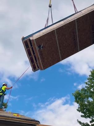 A crane dropping in part of the roof of a prefab home with a contractor helping guide the piece of roof.